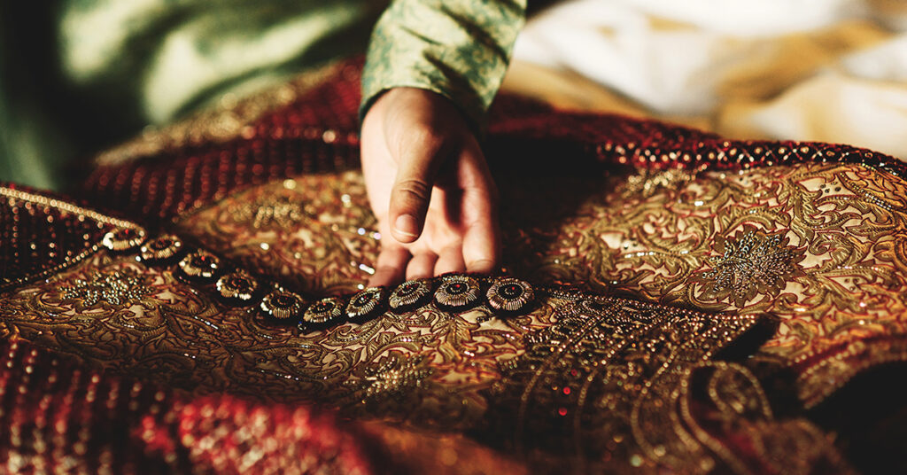 How to Choose Between a Sherwani and a Suit for Your Big Day? 1 Person's hand touching detailed, ornate embroidery on luxurious fabric.