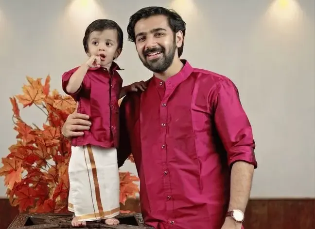 Adult and child in matching maroon shirts and white dhotis with maroon borders, bonding through traditional Indian attire, standing next to a decorative wooden stand with an orange plant in the background.
